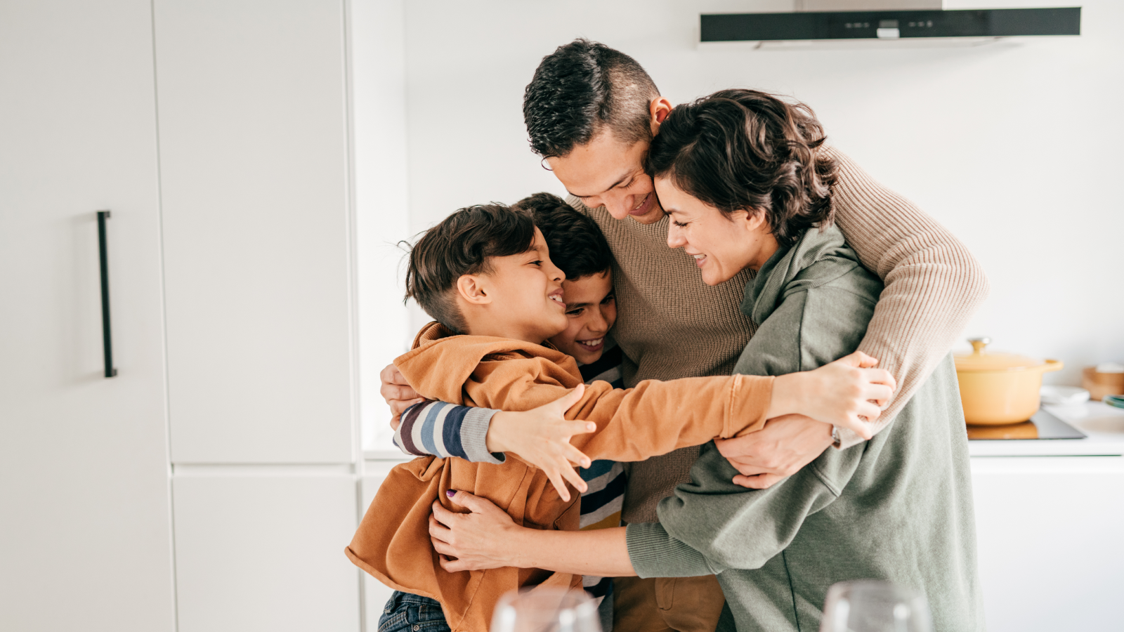 Family of four hugging and laughing in kitchen.